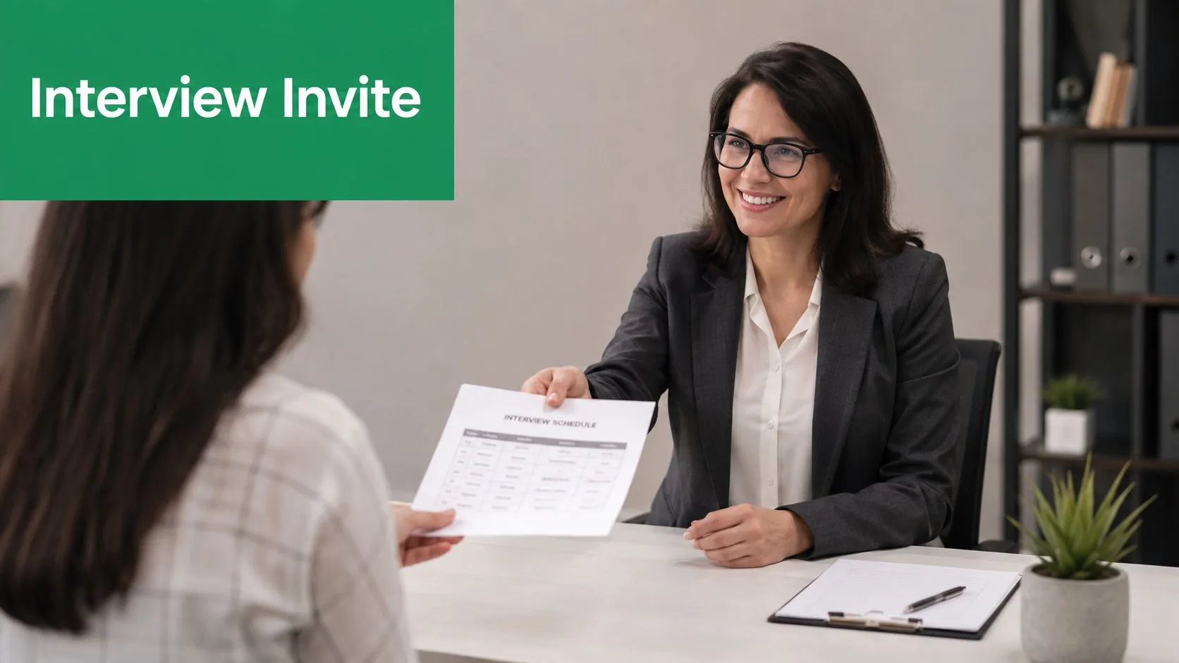 A smiling interviewer in a suit handing an interview schedule paper to a job candidate.